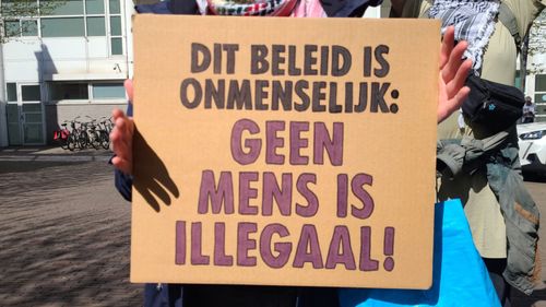 Persoon met bord met de tekst 'DIT BELEID IS ONMENSELIJK: GEEN MENS IS ILLEGAAL!'