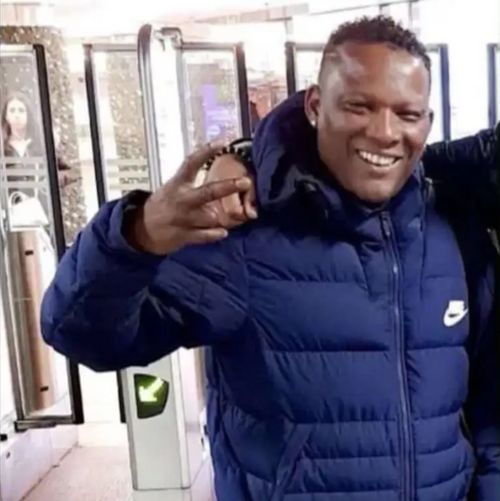 Foto van Alfredo Gulu. Hij is een donkere man met een blauwe winterjas, die lacht en een paar vingers opsteekt als een groet. In zijn andere hand heeft hij een telefoon vast. Een ander persoon slaat een arm om hem heen.